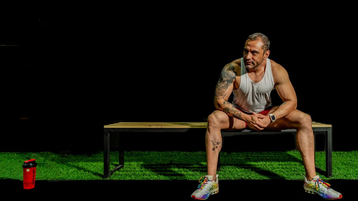 A muscular, tattooed man seated on a bench in a gym, depicting modern fitness lifestyle.